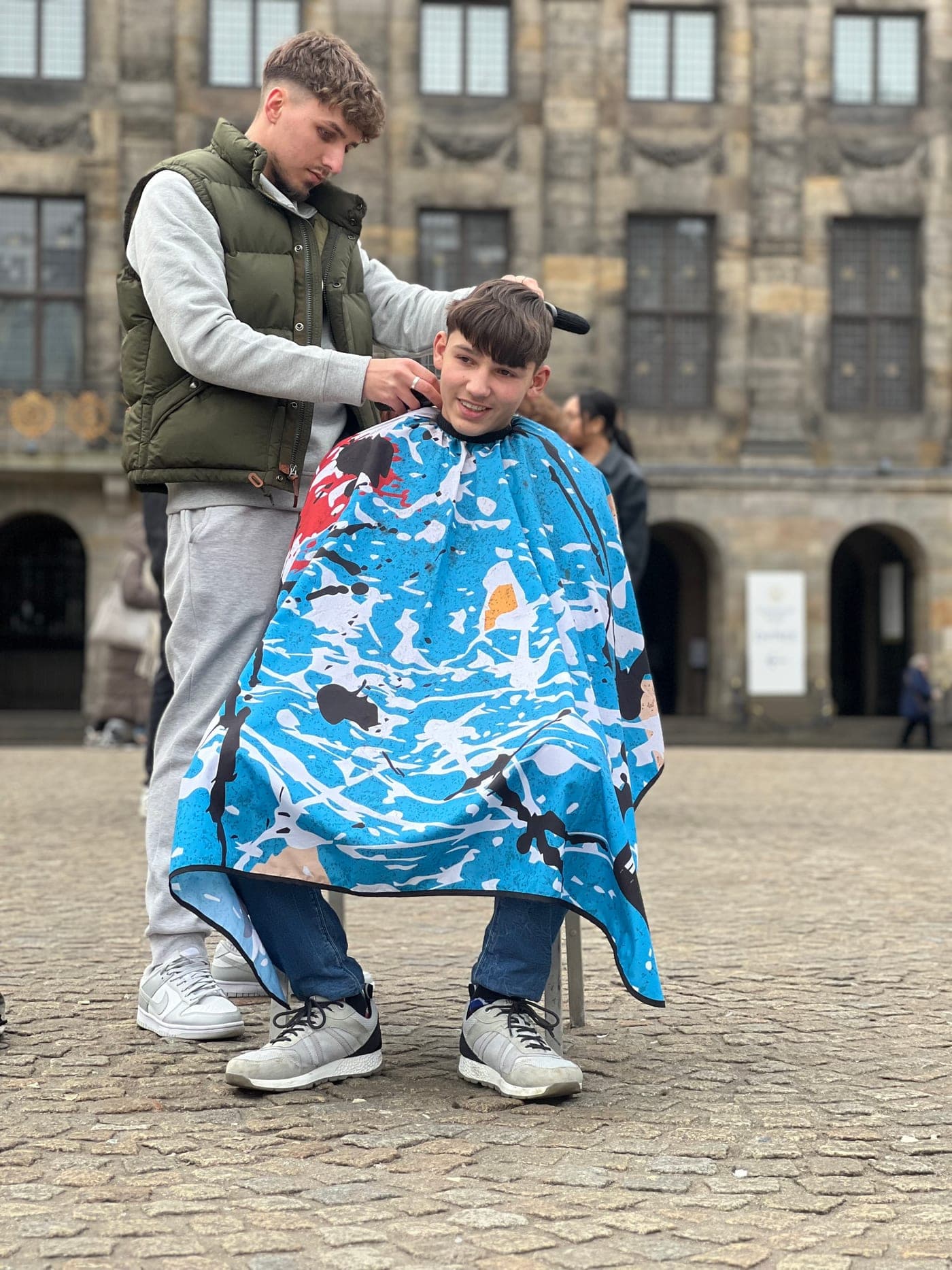 Krystian cutting a young client's hair outdoors in a city square in Amsterdam, in front of a grand old building.